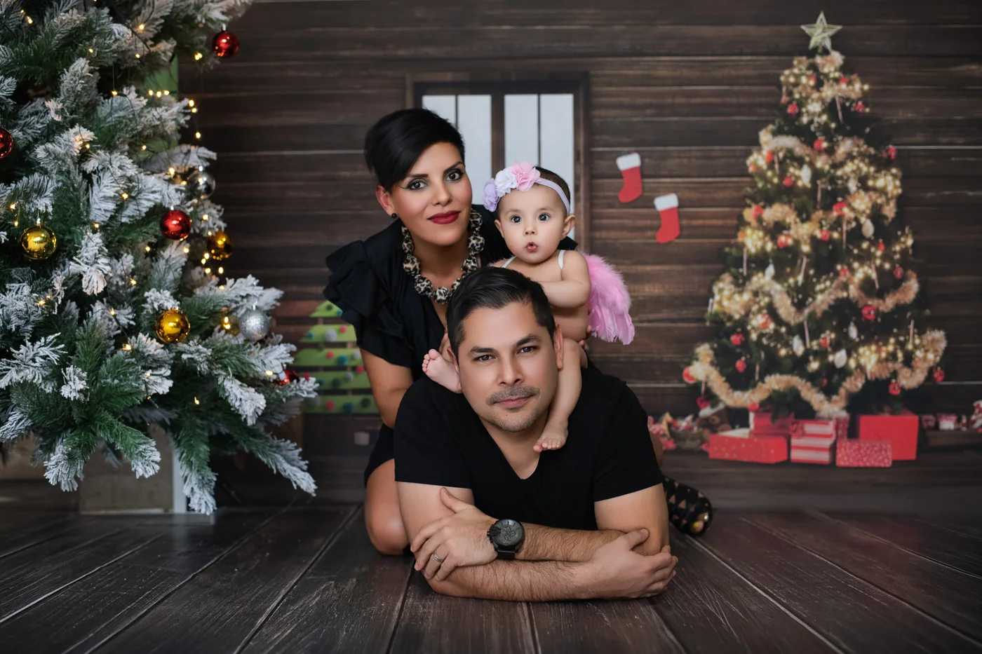Retrato familiar de dos adultos y un bebé, entre árboles de Navidad y un fondo de madera.